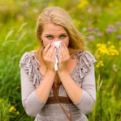 lady in tan dress holding a tissue to nose with both hands whilst standing in a meadow