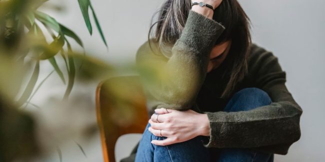 lady in jeans and jumper sat with knees up on wooden chair with arm around knees and hand on head