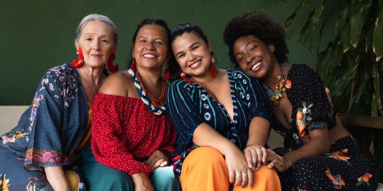 4 women all sat in a row on a sofa wearing colourful clothing
