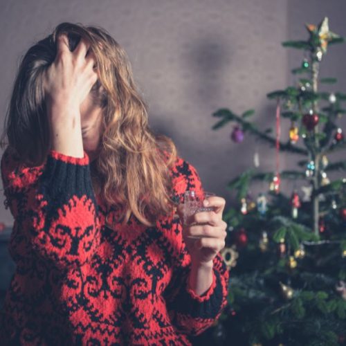 woman clutching a wine glass and her head in front of a christmas tree